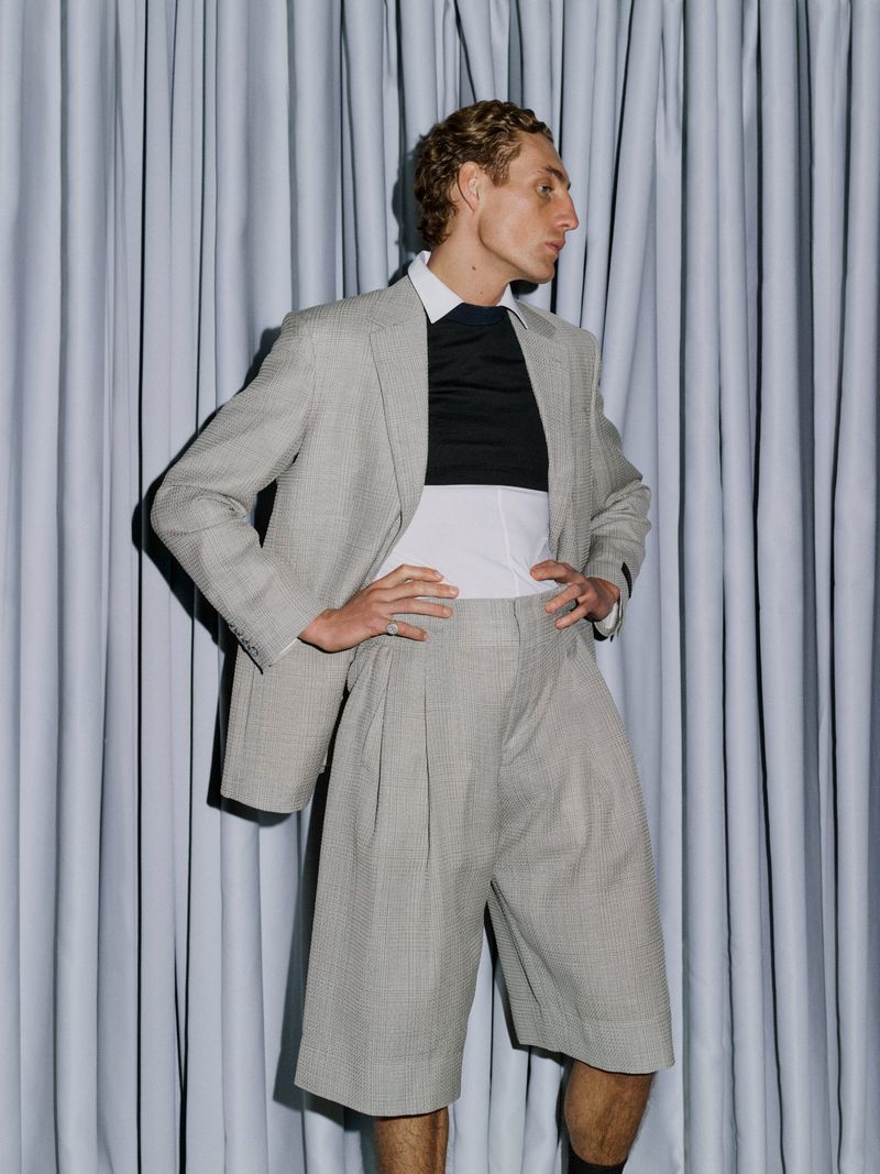 Model in a grey suit jacket and matching shorts standing against gathered grey fabric drapes in a fashion portrait.