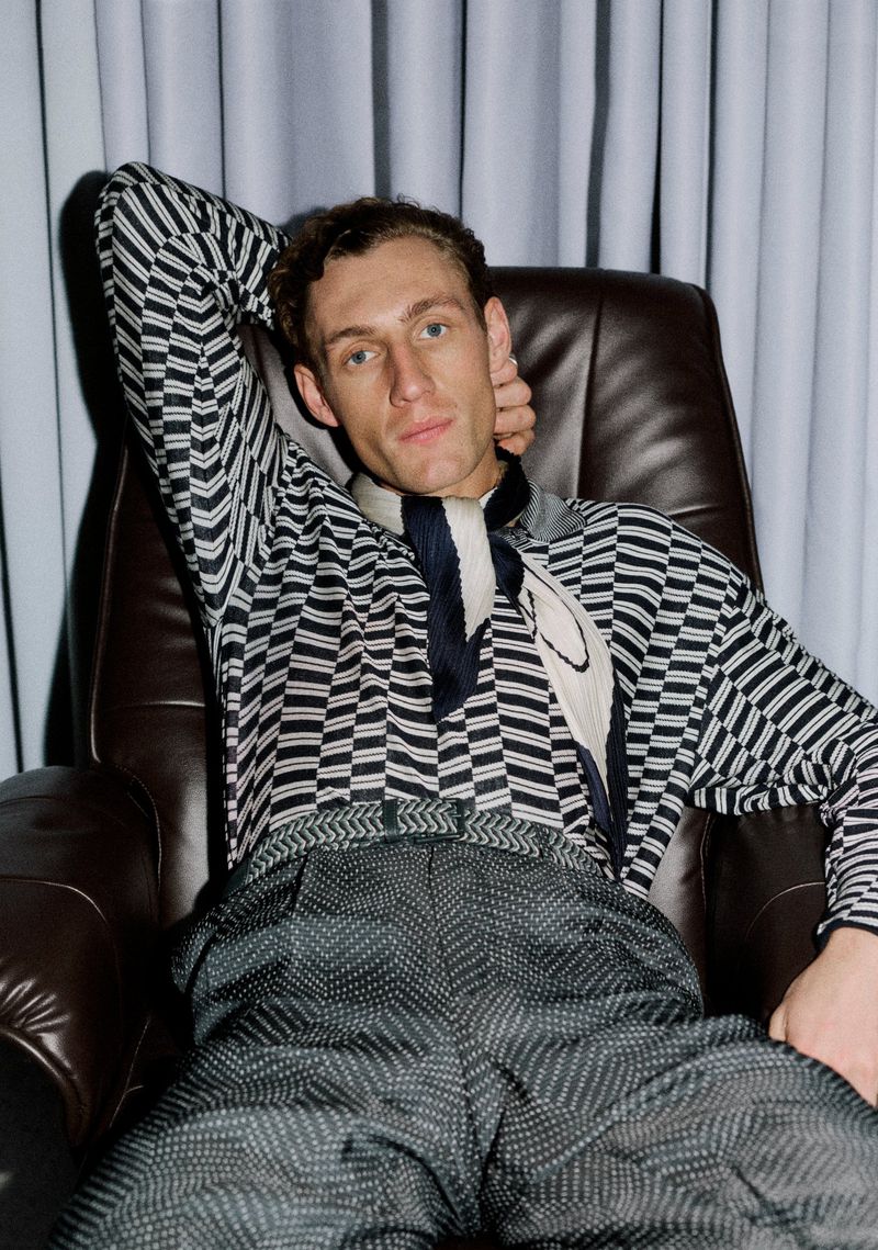 Aubrey O'Mahony seated in a brown leather armchair wearing a patterned knit shirt and neck scarf in front of grey curtains.