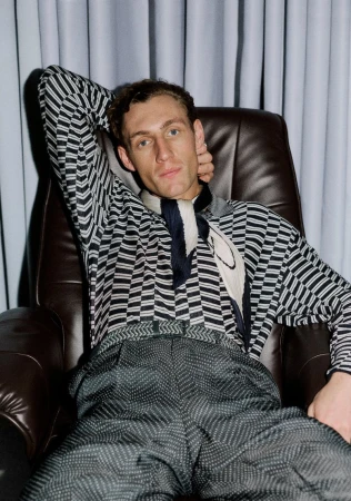 Aubrey O'Mahony seated in a brown leather armchair wearing a patterned knit shirt and neck scarf in front of grey curtains.