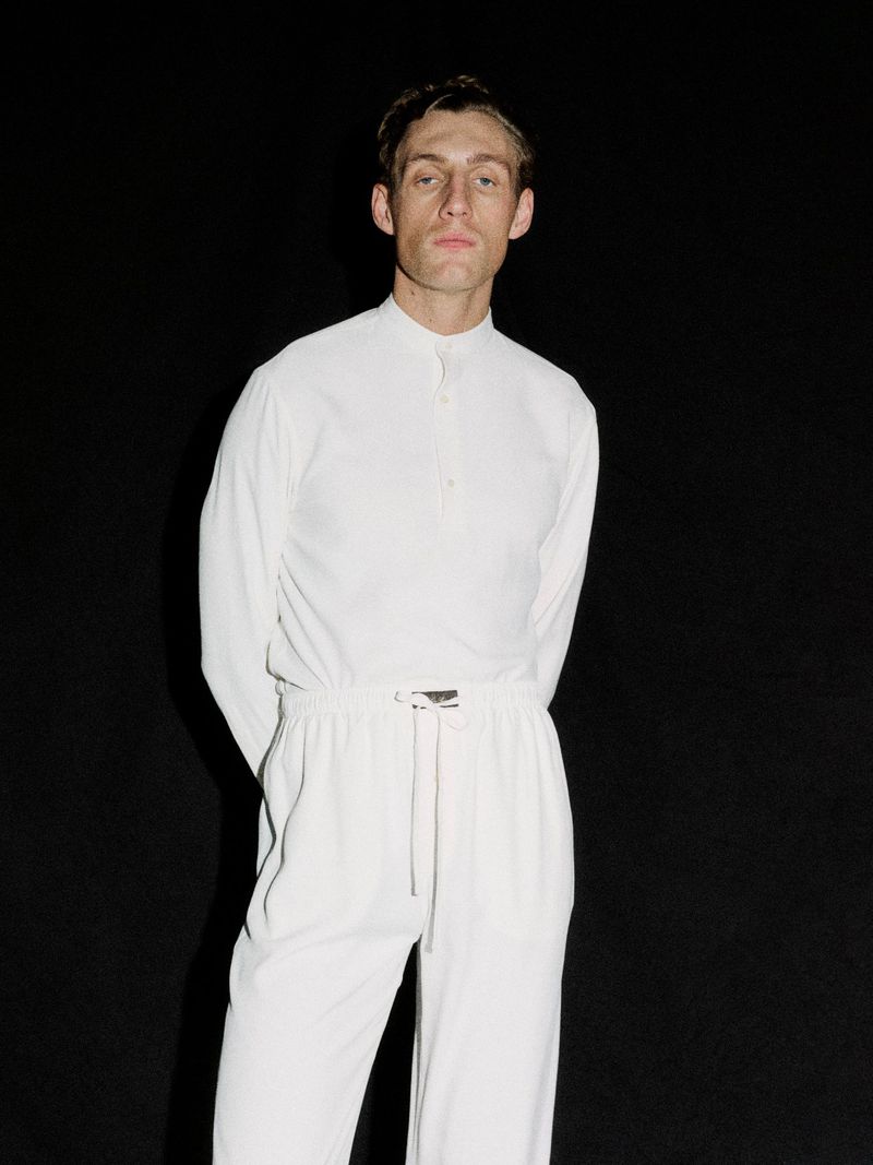 Model Aubrey O'Mahony wearing a white long-sleeve shirt and drawstring trousers against a solid black background.