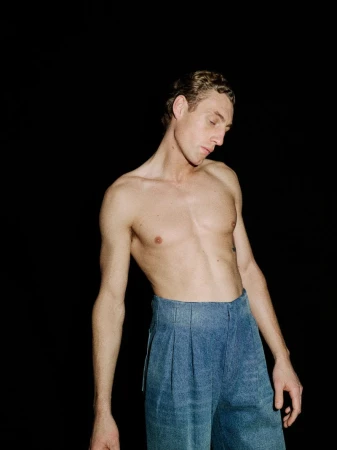 Shirtless male model wearing blue high-waisted trousers against a solid black background
