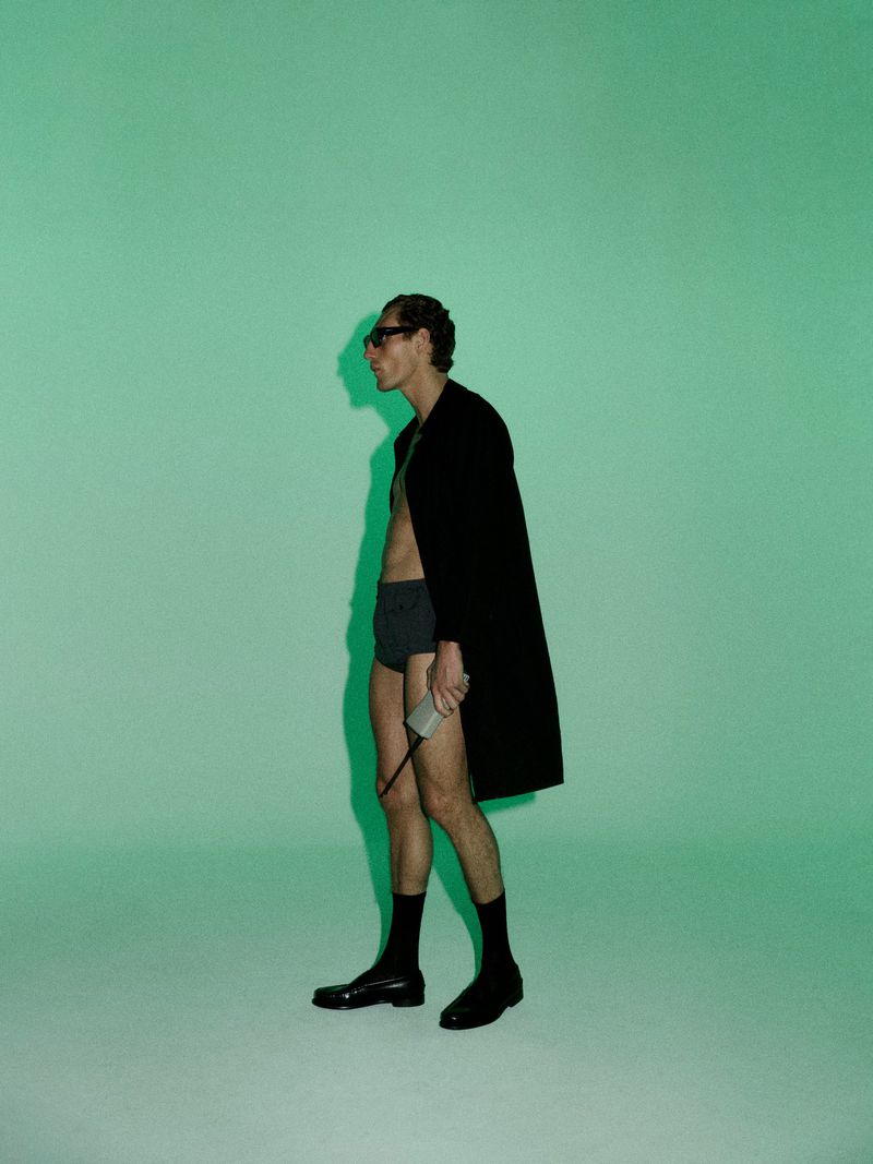 Aubrey O'Mahony wearing an open black coat, dark briefs, and sunglasses against a mint green backdrop.
