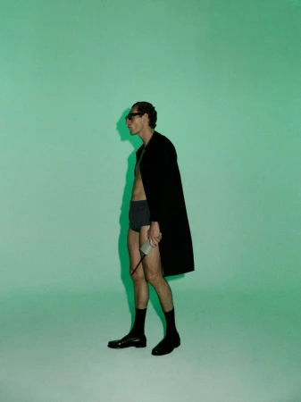 Aubrey O'Mahony wearing an open black coat, dark briefs, and sunglasses against a mint green backdrop.