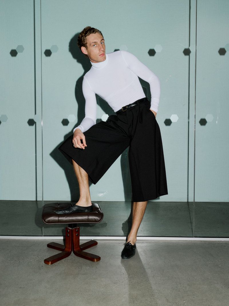 Aubrey O'Mahony in a white turtleneck and black culottes with one foot on a leather stool against glass panels.