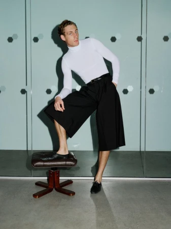 Aubrey O'Mahony in a white turtleneck and black culottes with one foot on a leather stool against glass panels.