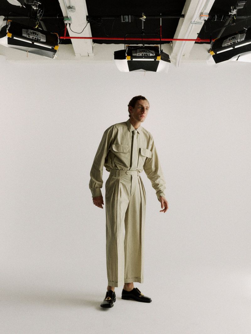 Model Aubrey O'Mahony stands in a matching beige shirt, tie, and pleated trousers in a photo studio beneath overhead lighting.
