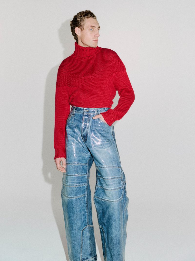 Aubrey O'Mahony poses in a red knit turtleneck sweater and wide-leg glossy blue jeans against a pale seamless studio backdrop.