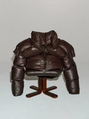 Dark brown leather puffer jacket styled over a mid-century swivel armchair against a pale grey studio backdrop.