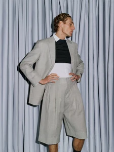 Model in a grey suit jacket and matching shorts standing against gathered grey fabric drapes in a fashion portrait.