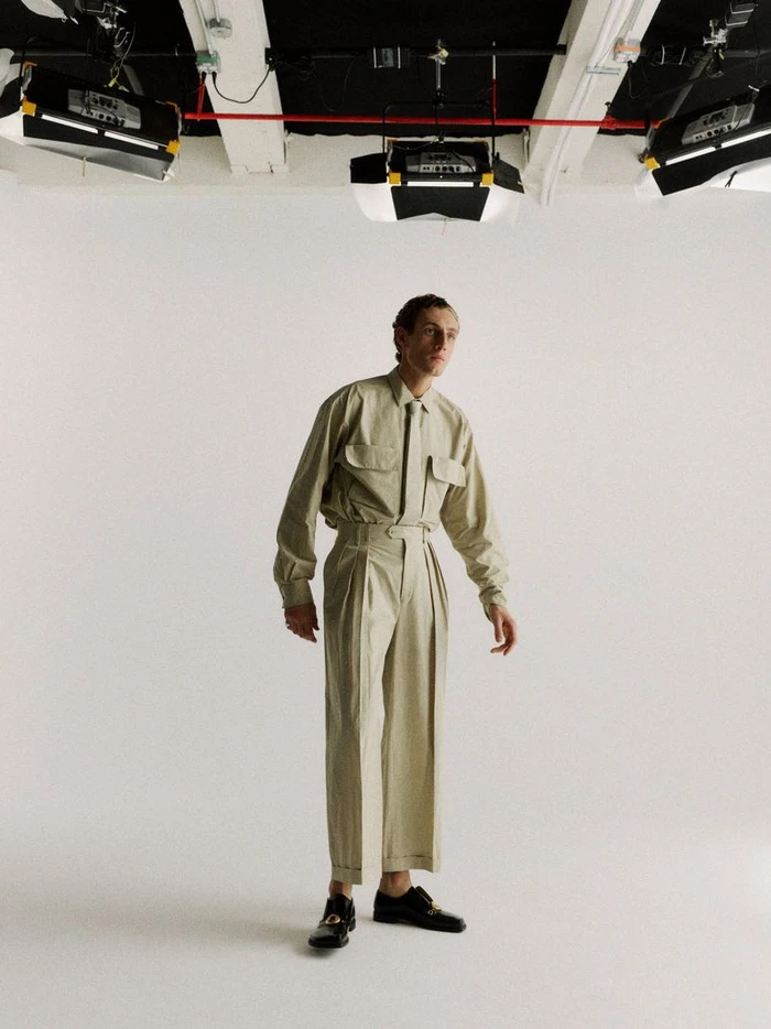 Model Aubrey O'Mahony stands in a matching beige shirt, tie, and pleated trousers in a photo studio beneath overhead lighting.