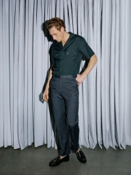 Model Aubrey O'Mahony standing in a dark short-sleeve shirt, pleated trousers, and loafers in front of a grey curtain.