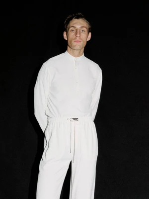 Model Aubrey O'Mahony wearing a white long-sleeve shirt and drawstring trousers against a solid black background.