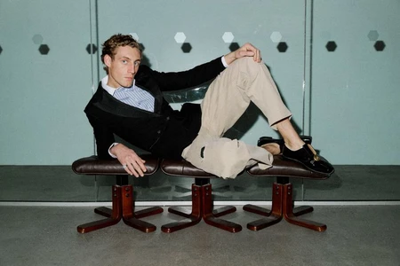 Model Aubrey O'Mahony lounges across three brown leather swivel stools wearing a black blazer, beige trousers, and loafers.
