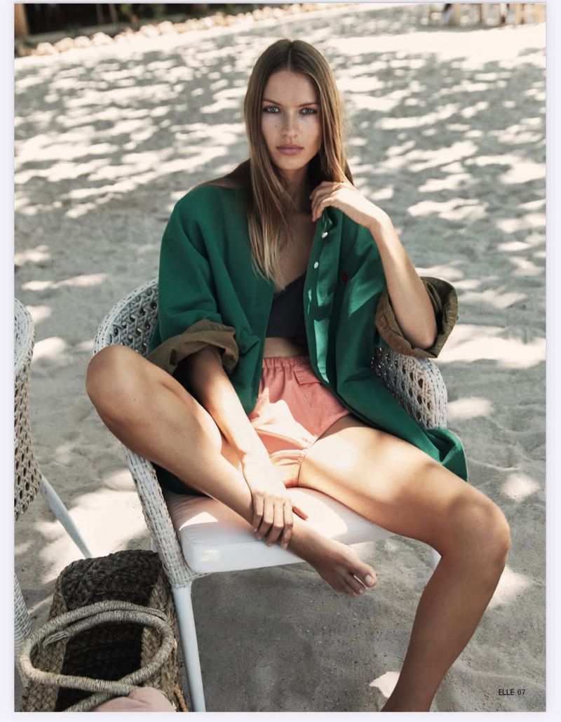 Model Lea Seelenmeyer seated in a wicker chair on a sandy beach wearing an oversized green overshirt and peach shorts.