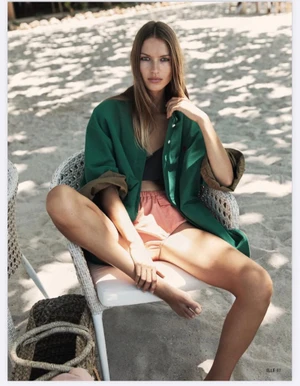 Model Lea Seelenmeyer seated in a wicker chair on a sandy beach wearing an oversized green overshirt and peach shorts.