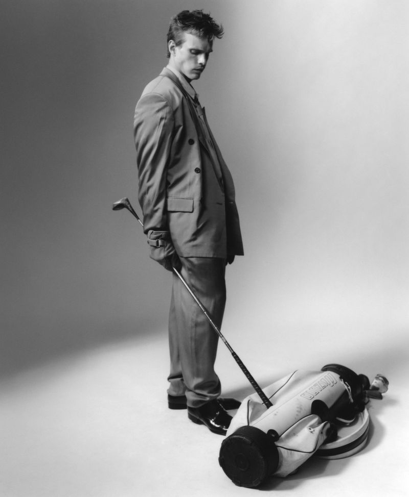 Finn Bennett wearing a double-breasted suit holding a golf club beside a golf bag on a studio floor