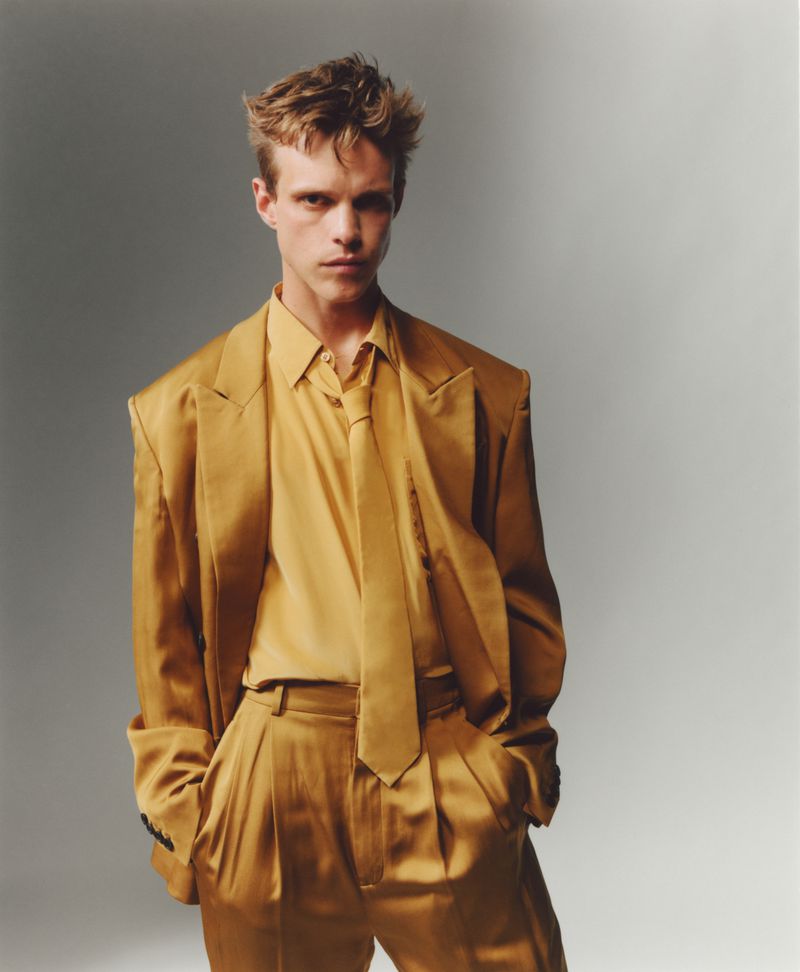 Finn Bennett wearing a monochromatic golden-yellow satin suit, matching shirt, and tie against a neutral grey studio background.