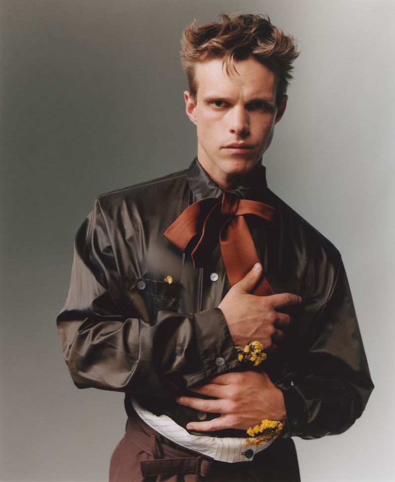 Actor Finn Bennett posing in a dark brown satin shirt with a large neck bow and yellow floral accents against a grey studio backdrop.