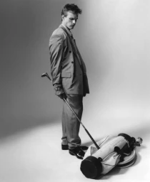 Finn Bennett wearing a double-breasted suit holding a golf club beside a golf bag on a studio floor
