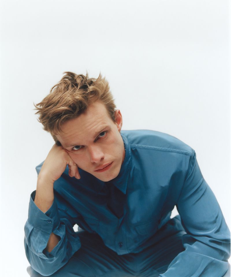 Actor Finn Bennett resting his head on his hand while wearing a blue button-down shirt against a white studio background.