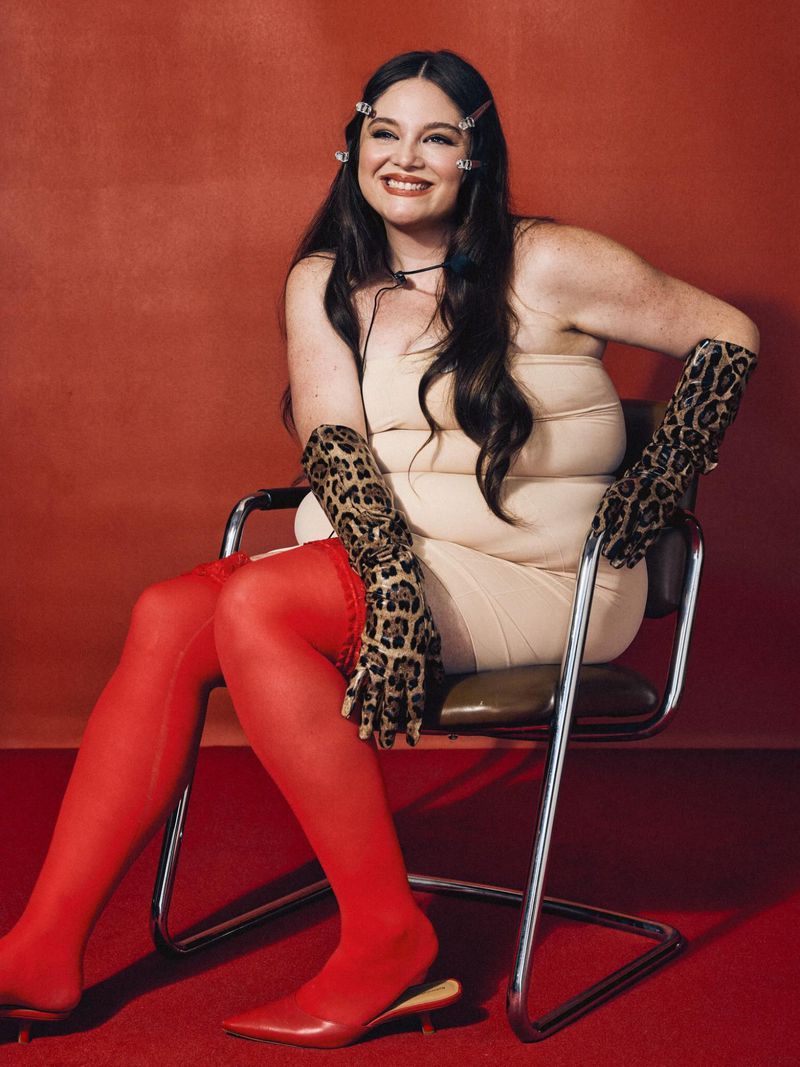 Meg Stalter seated in a chrome chair wearing a nude strapless dress, leopard-print gloves, and vibrant red tights against a red wall.