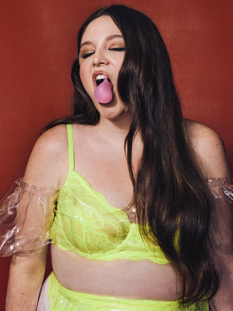 Meg Stalter posing in a neon yellow lace bralette with a pink bubble in her mouth against a textured reddish-brown background.