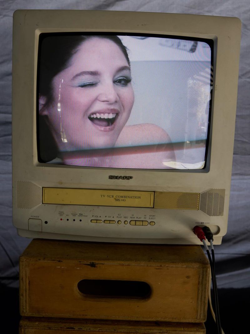 Vintage Sharp CRT television on a wooden crate displaying actress Meg Stalter winking and smiling on screen.