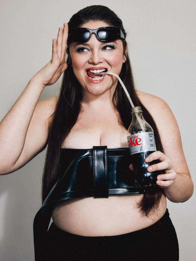 Meg Stalter wearing black sunglasses and a patent leather corset top while holding a glass soda bottle with a straw against a grey background.