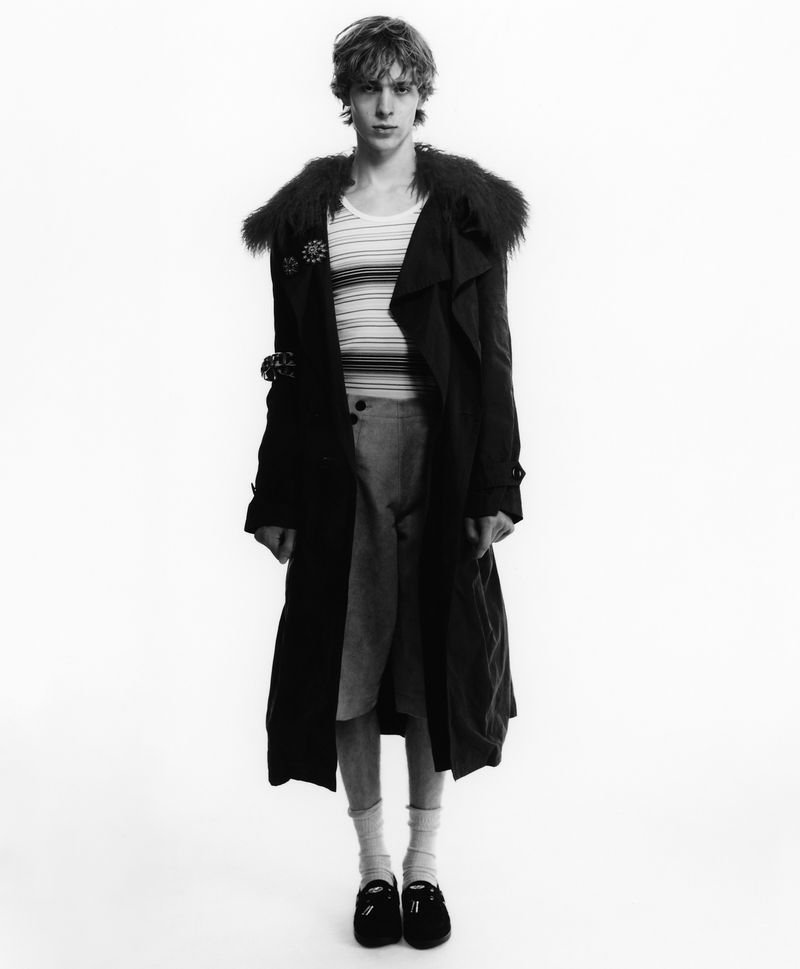 Male model in a black fur-trimmed coat, striped top, and shorts standing against a pure white studio backdrop.
