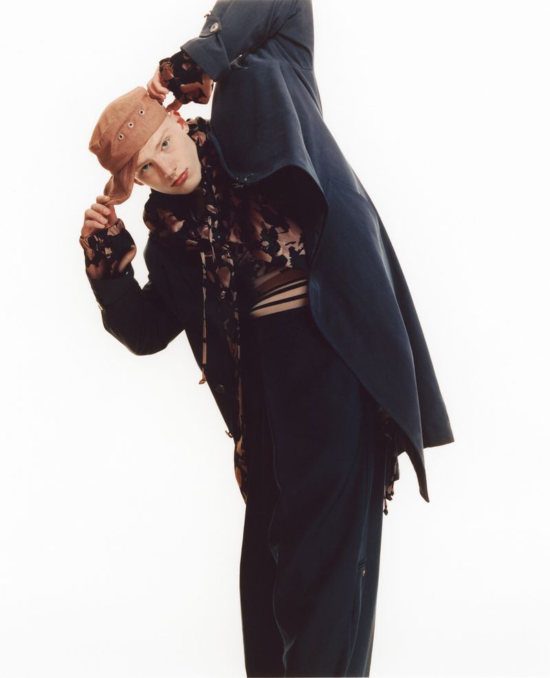 A male model wearing an oversized dark navy coat and a brown cap poses against a plain white studio background.