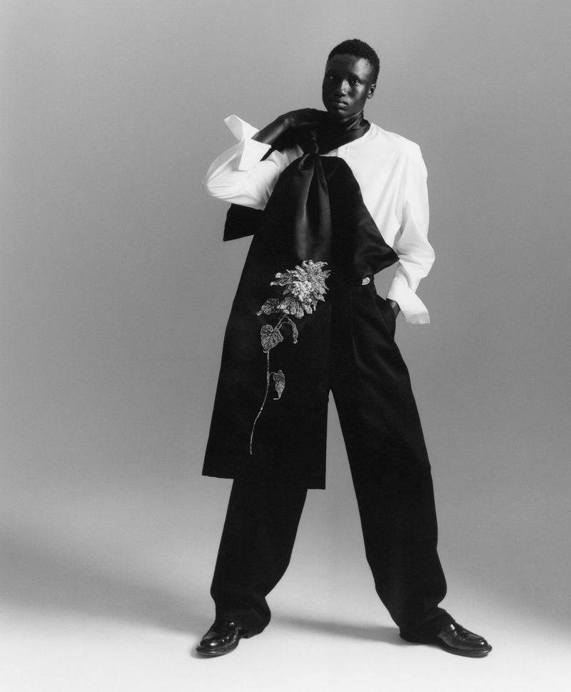 Model Jum Kuochnin posing in a white shirt, wide-leg black trousers, and an embroidered black drape against a studio backdrop.