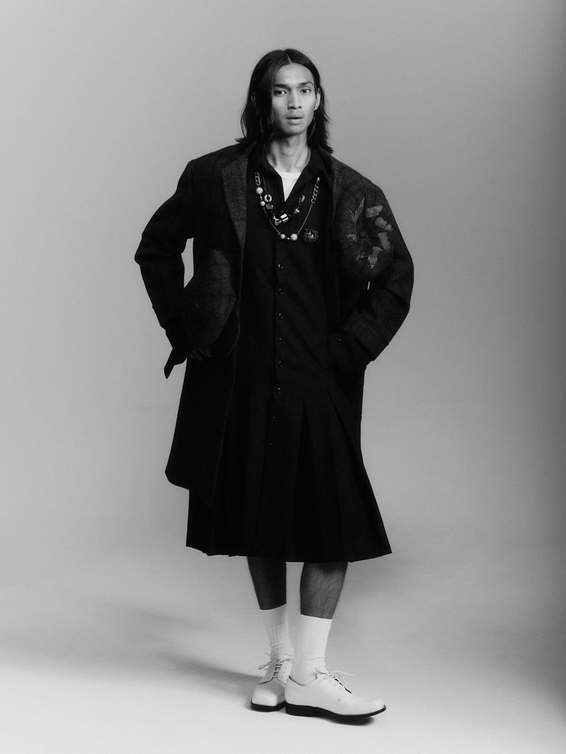Model Ridzman Zidaine wearing a long black coat, pleated skirt, and white brogues against a studio backdrop.