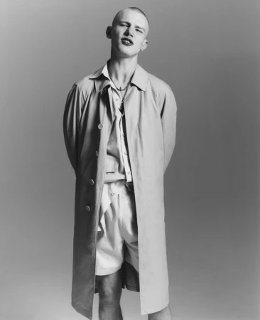 Black and white portrait of a male model with a buzz cut wearing an open trench coat and shorts in a studio setting.