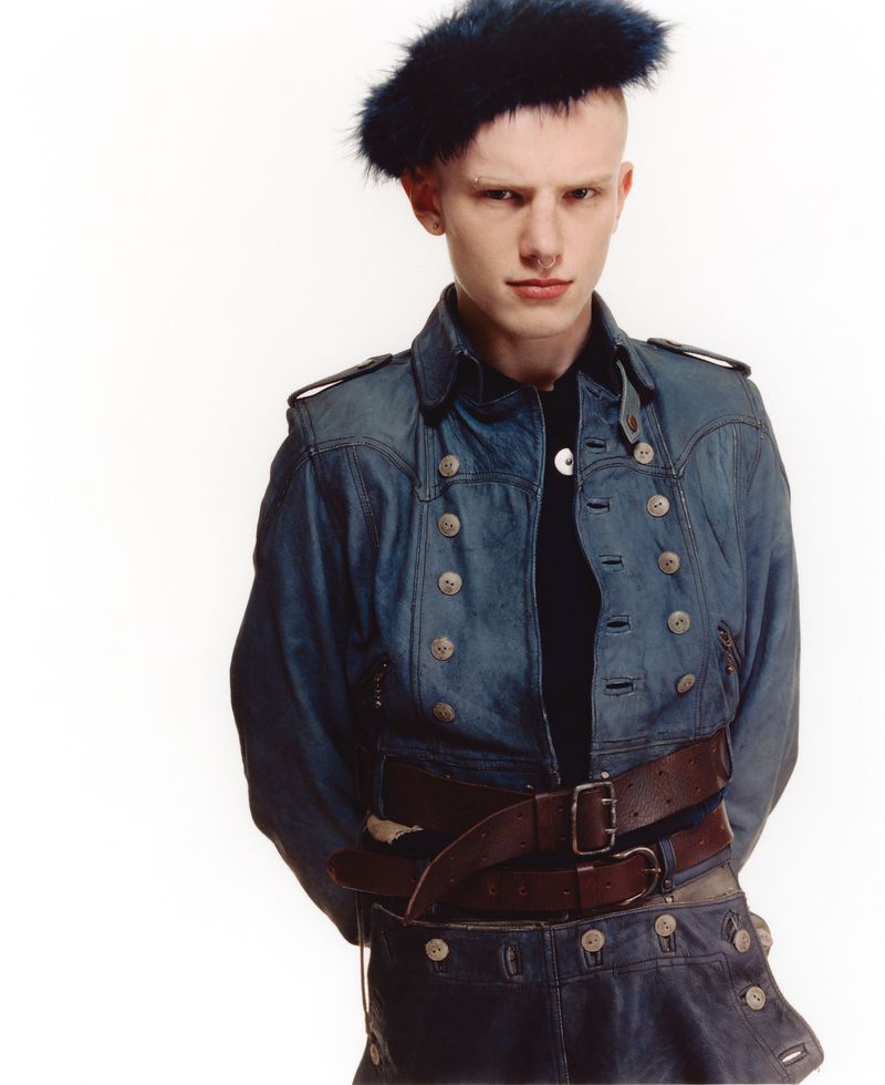 Model wearing a blue denim military jacket, layered brown leather belts, and a fuzzy navy beret against a plain white background.