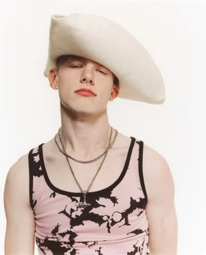 A young man wearing an oversized cream felt beret and a pink patterned tank top against a plain white studio backdrop.
