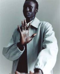 Fashion portrait of a Black male model extending his hand forward while wearing a perforated light grey jacket against a studio backdrop.
