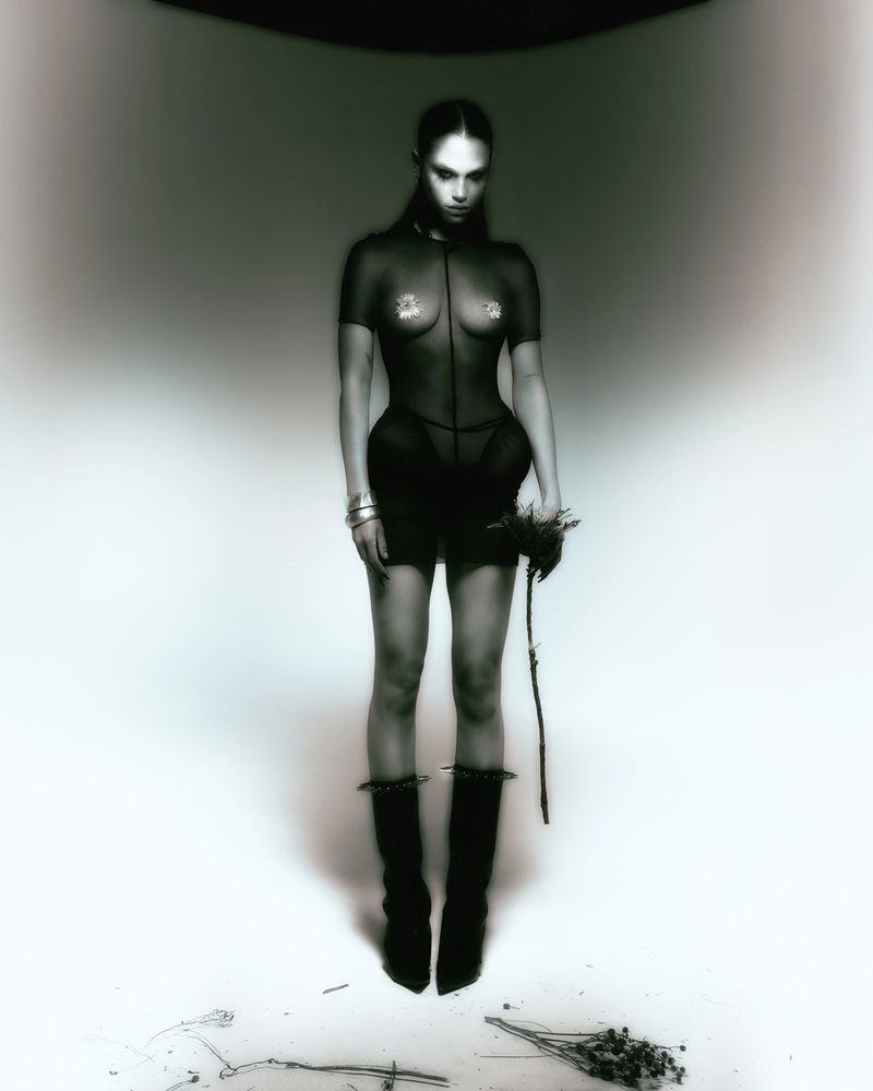 Model Meredith Mickelson posing in a sheer black corseted bodysuit and black boots against a studio backdrop.