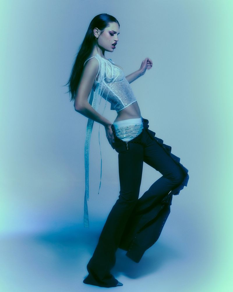 Model Meredith Mickelson posing in a sheer white sleeveless top and black trousers against a cool blue studio backdrop.