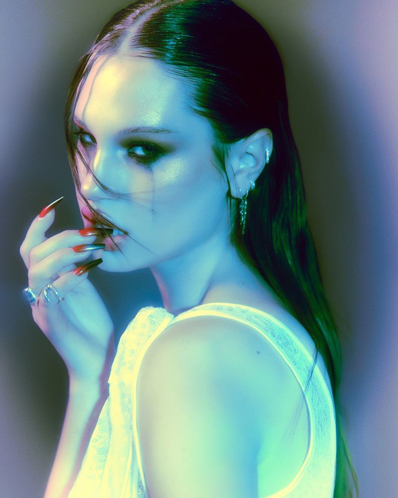 Meredith Mickelson touching her lip with long stiletto ombre nails, styled in smoky eye makeup and a lace top under cyan lighting.