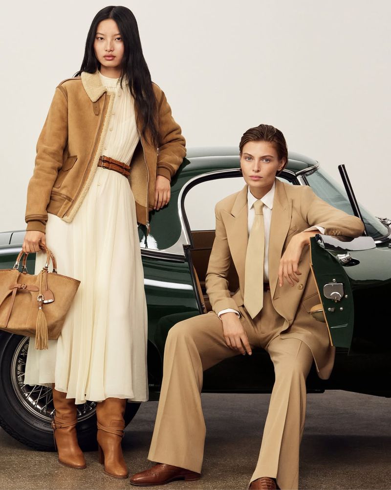 Two women model vintage fashion next to a vintage green car. One woman sits on the car door in a tan suit, the other stands with a purse.