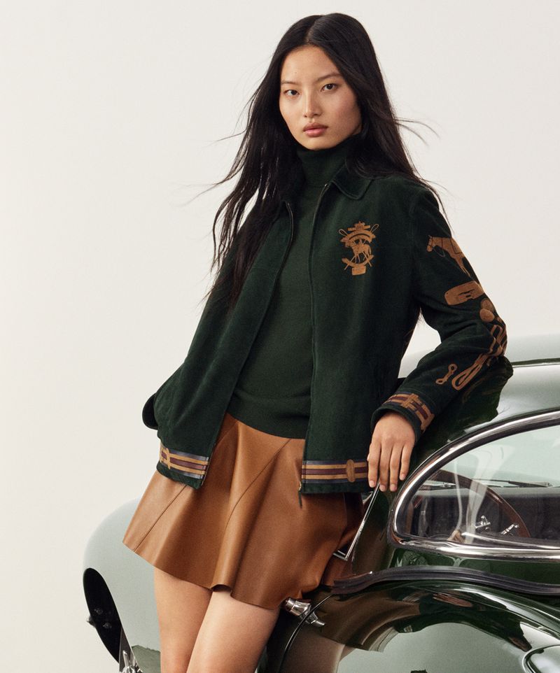 Brunette woman in a dark green jacket and brown mini skirt poses leaning on the dark green hood of a vintage car indoors.