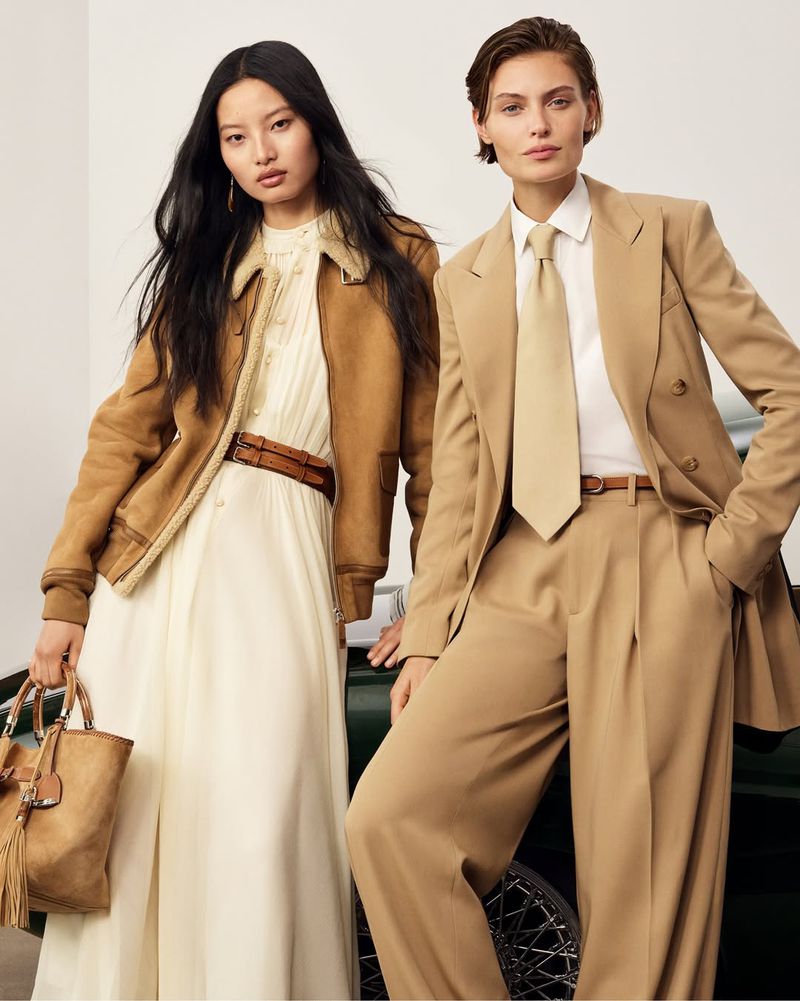 Two fashion models in tan suits and jacket pose near a green vintage car, showcasing complementary neutral-toned outfits and a handbag.