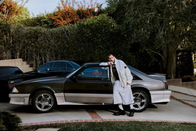 Man in an oversized light jacket and matching long shorts leaning against a parked grey sports car on a residential driveway.