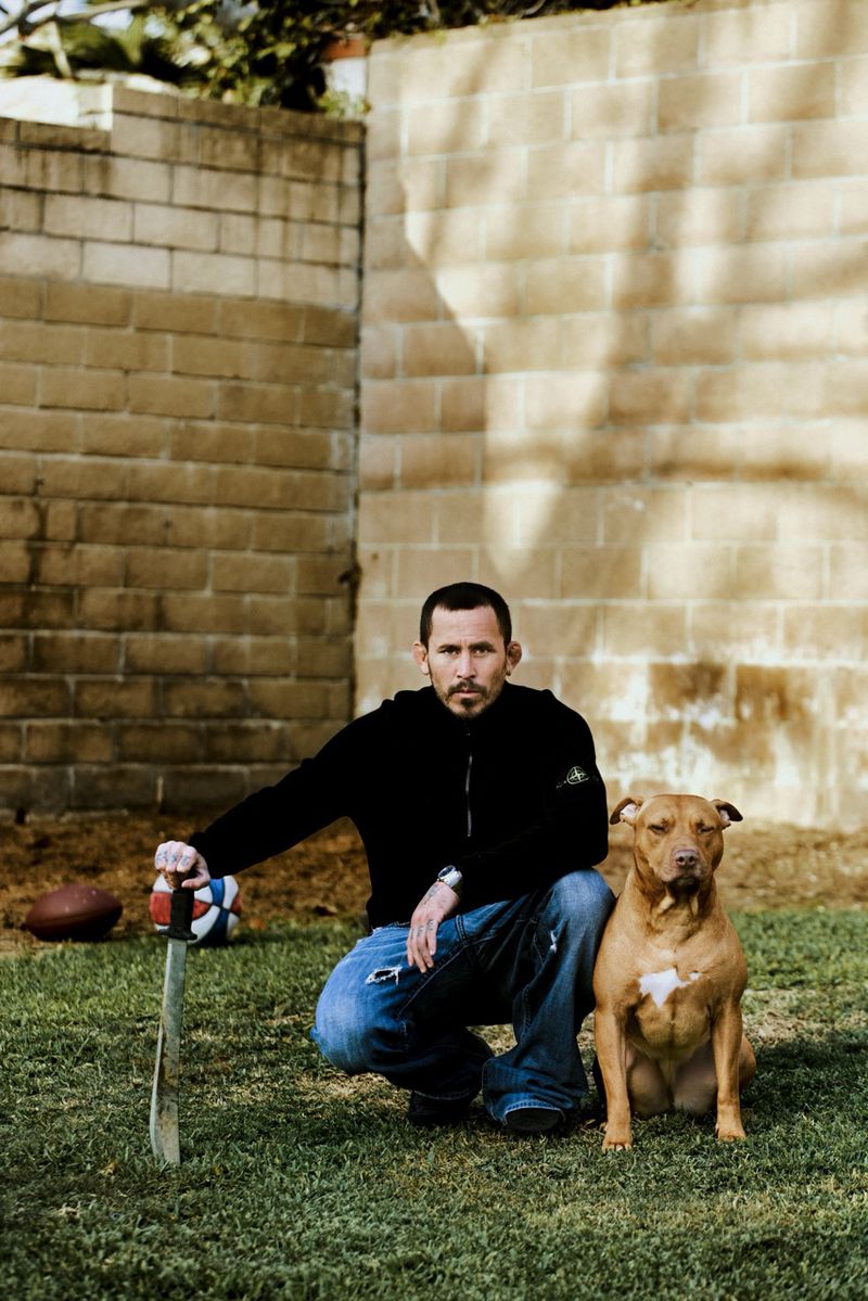 Man crouching on a grass lawn holding a machete beside a brown pit bull in front of a tan cinder block wall.