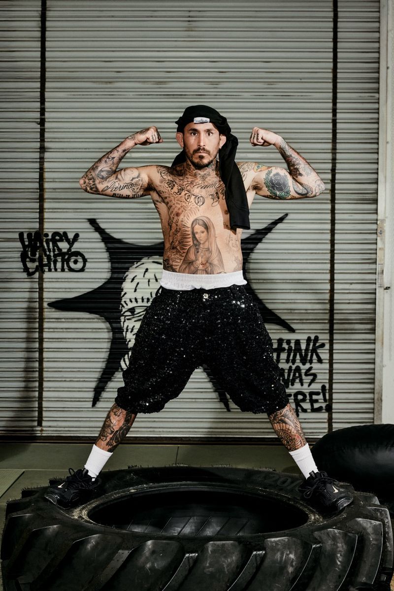 Tattooed man flexing muscles wearing sequin shorts and standing inside a large tire for an i-D Magazine editorial.