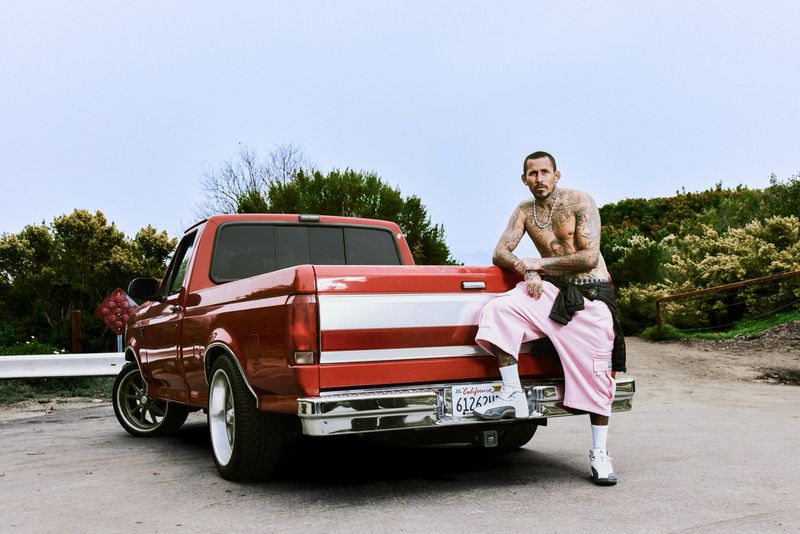 Tattooed man leaning against the tailgate of a red pickup truck wearing pink shorts and sneakers