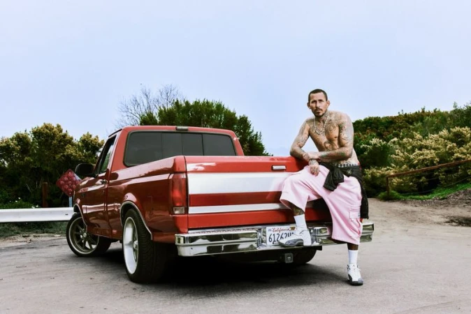 Tattooed man leaning against the tailgate of a red pickup truck wearing pink shorts and sneakers