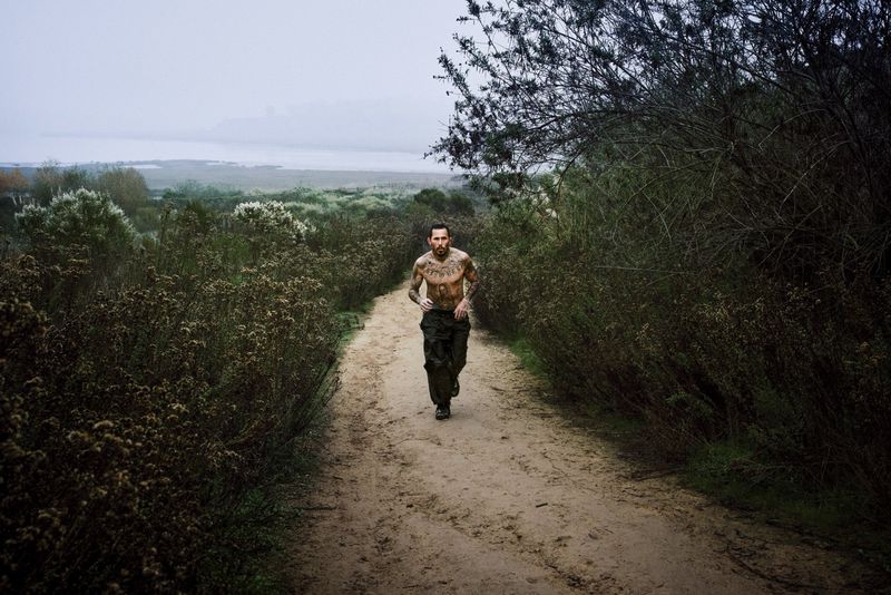 A shirtless tattooed man runs down a sandy dirt trail surrounded by dense wild brush under an overcast sky.