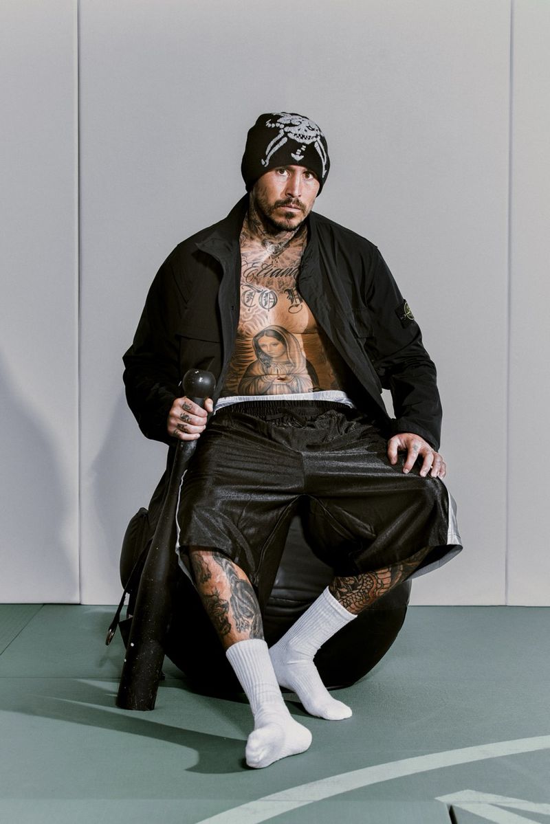 Tattooed man wearing a black beanie and jacket holding a club while seated for i-D Magazine