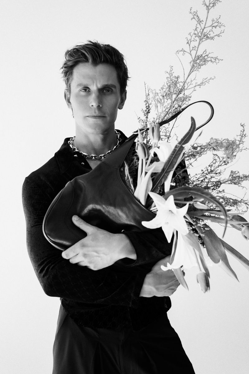 Antoni Porowski wearing a dark shirt and chain necklace, holding a black leather bag filled with lilies against a white backdrop.
